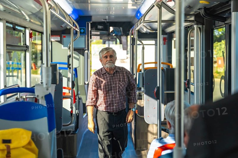 Senior man with a beard and plaid shirt standing in the aisle of a public bus, commuting during the day, with other passengers seated and windows bringing in natural light