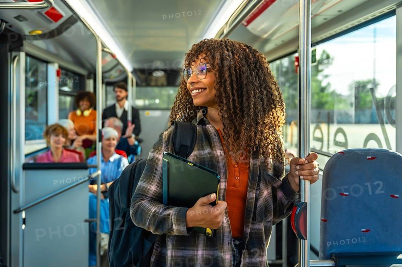 Young black woman with curly hair and glasses smiling, holding a digital tablet and a pole while standing in a public bus filled with diverse passengers, representing urban travel and daily commute