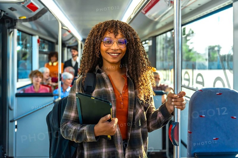 Young black woman student smiling at the camera while standing inside a public transportation bus, holding her backpack and tablet, traveling during daytime