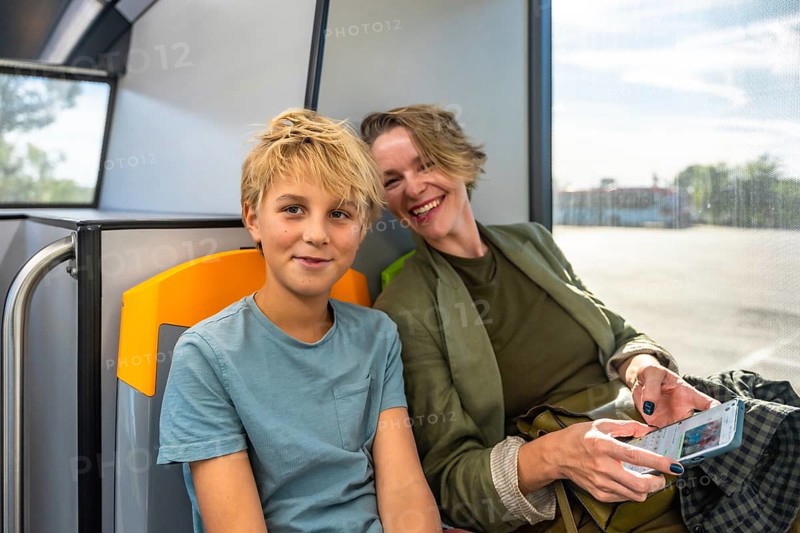 Mother and son sitting inside a bus, sharing a moment while traveling using public transport, the mother scrolling on a smartphone and the son looking at the viewer