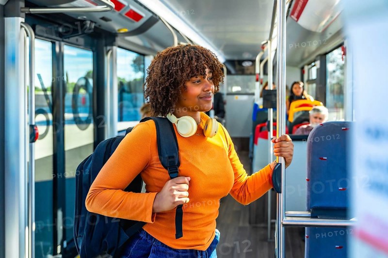 Young black woman with headphones and backpack standing in a public transport bus, holding a handrail and smiling, representing urban lifestyle and daily commute