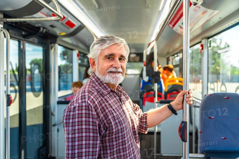 Senior man with a beard and plaid shirt smiling at the camera while holding a pole inside a public transport bus, representing urban life and daily commutes