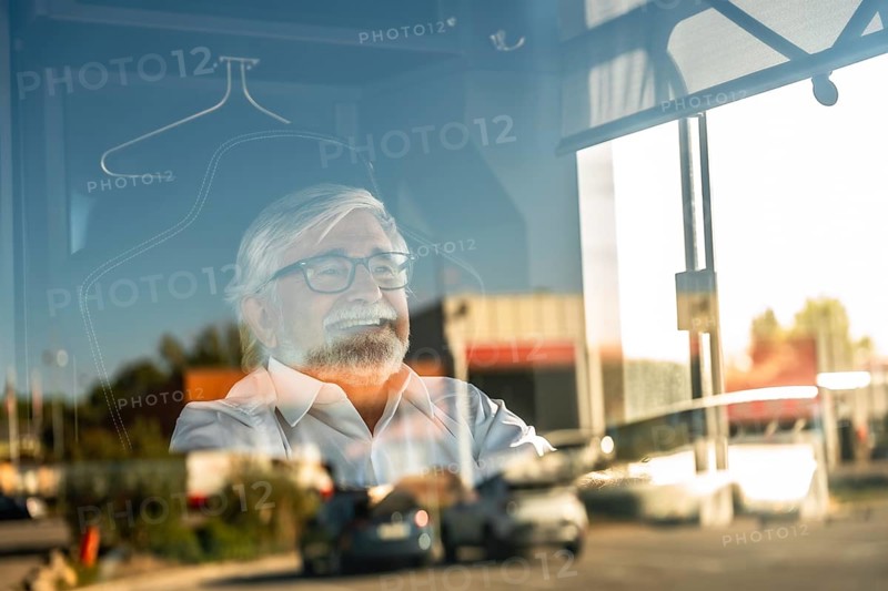 Senior bus driver smiling while looking outside through a window, reflecting street traffic, traveling and enjoying a journey, showing happiness and positive emotions during work