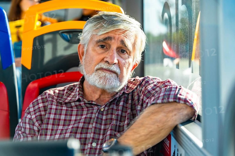 Senior man with white beard and plaid shirt sits by a bus window, gazing pensively at the camera during an urban commute, conveying quiet reflection and daily routine