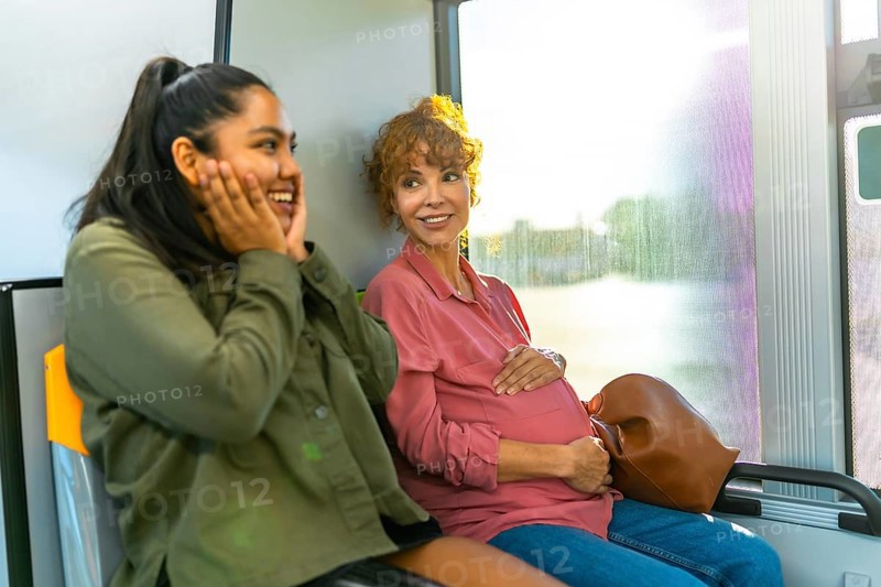 Young woman is smiling and showing surprise while looking at her pregnant friend, who is gently cradling her baby bump, both traveling together on a public bus