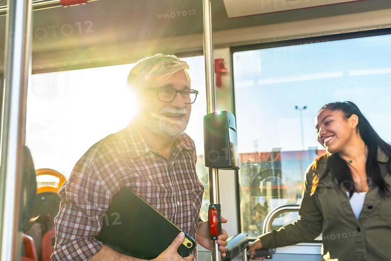 Two smiling passengers, an older man and a young woman, are traveling on a bus during daylight, experiencing a pleasant journey using public transportation