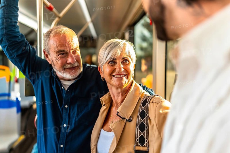 Happy senior couple standing on a city bus, smiling and chatting with another passenger while holding the handrail, enjoying a bright, relaxed commute together