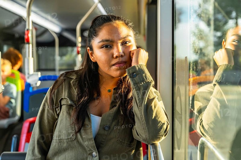Young woman riding public transport contemplating and looking at camera while warm sunlight reflects on the bus window, creating a thoughtful atmosphere