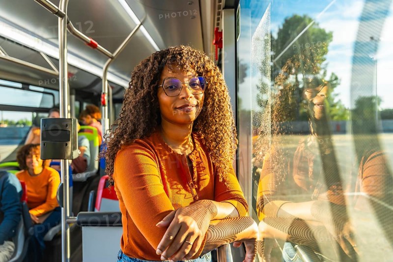 Smiling black woman with curly hair and glasses stands on a city bus during her commute, confident and happy, embodying urban mobility, independence and everyday city life