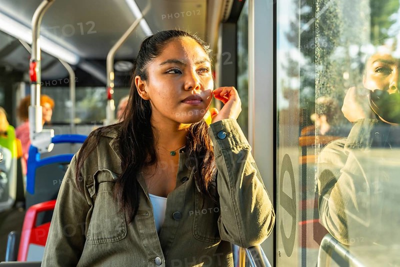 Young woman passenger traveling on a public transport bus, looking out of the window with a pensive expression, enjoying sunlight with her face reflecting on the glass