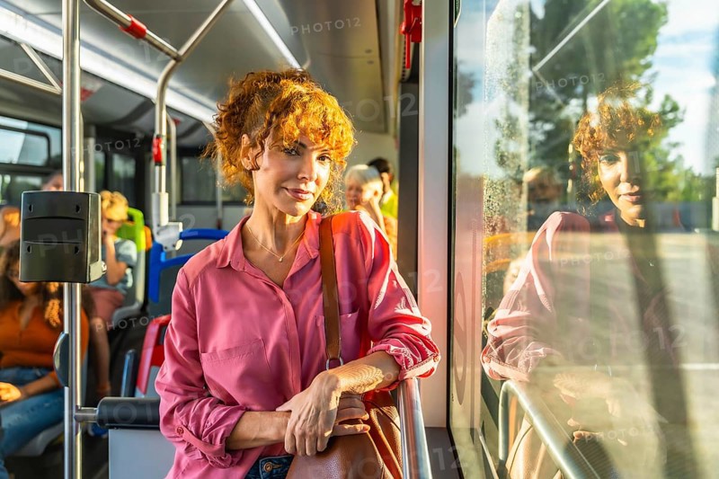 Young redheaded woman smiling and looking at camera while standing on a city bus, holding a bag amid other passengers during a sunny urban commute, casual and confident
