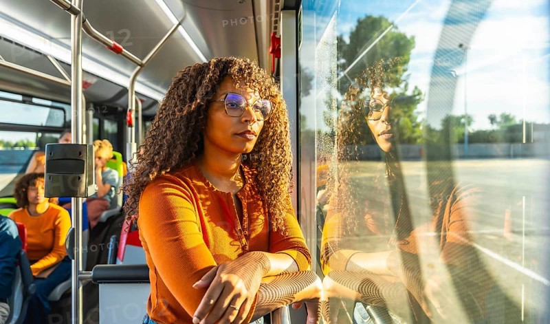 Young adult woman with curly hair and glasses looking out a public transport bus window, reflecting on her journey and commuting experience, with a thoughtful expression