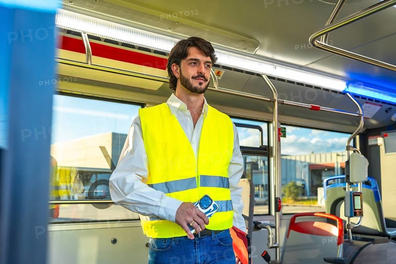 Man wearing an unbuttoned white shirt, blue jeans, and a yellow reflective safety vest, holding a phone, and standing confidently in a public transport bus