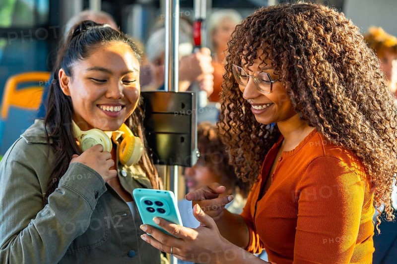 Two smiling multiethnic women bonding and sharing content on a smartphone while commuting together on a public transport bus, enjoying a pleasant journey