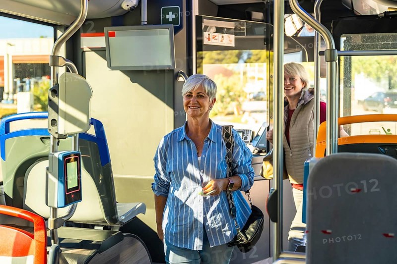 Smiling senior woman standing in a modern public transport bus, enjoying her journey and commuting using accessible urban transportation, with another passenger in the background
