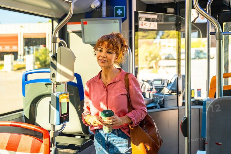 Woman standing on a city bus during the morning commute, holding a reusable coffee cup and brown shoulder bag, smiling and looking forward while traveling through an urban route