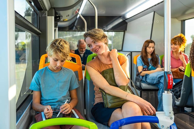 Mother and son share a city bus ride as the boy uses a tablet with a stylus, mother smiling nearby, bright interior, casual commute moments of learning and connection