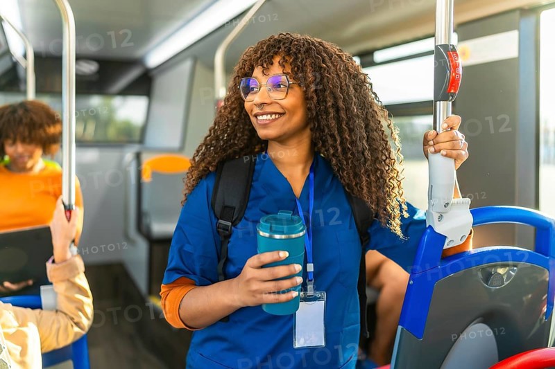 Young black nurse in uniform and backpack stands smiling on a busy city bus, holding a reusable cup as she commutes to work, eco conscious and ready for the day