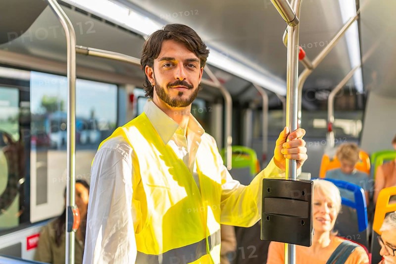 Young man in a high visibility safety vest stands on a modern city bus, holding a pole while commuting, smiling confidently among fellow passengers on board