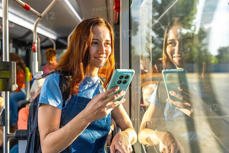 Young woman smiling on a sunny bus commute, holding her smartphone and looking out the window with her reflection in the glass, relaxed and connected during travel