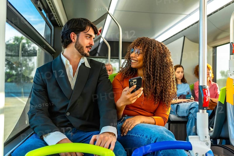 Diverse couple traveling on a public bus, the woman showing content on her smartphone to the man while they are talking and smiling, enjoying their daily commute together