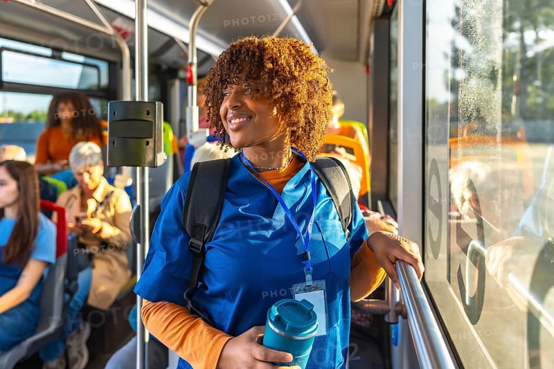 Smiling young black nursing student in blue scrubs with backpack and reusable cup, standing on a city bus and looking out the window during her commute to clinicals