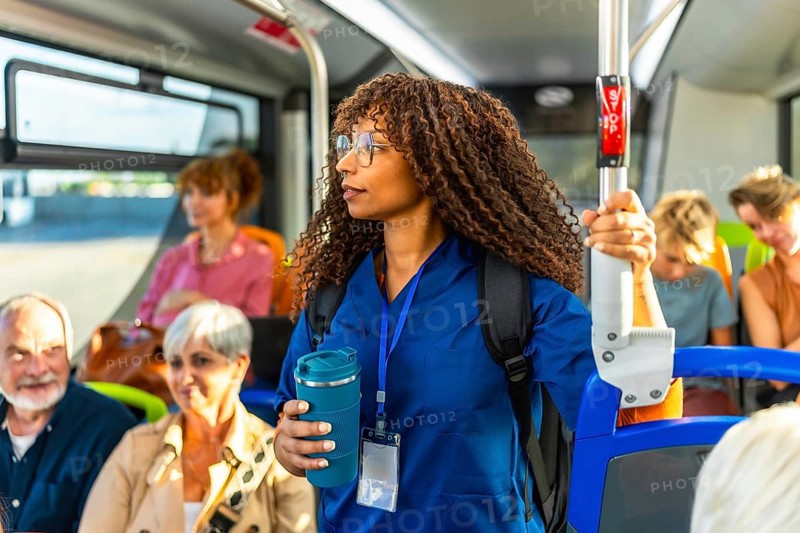 Young woman nurse in medical scrubs carrying a backpack and holding a reusable coffee cup, traveling on a crowded public transport bus filled with diverse passengers