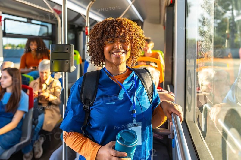 Young black woman doctor or student nurse standing on a public transport bus, wearing blue scrubs, a backpack, and carrying a travel mug, smiling at the camera