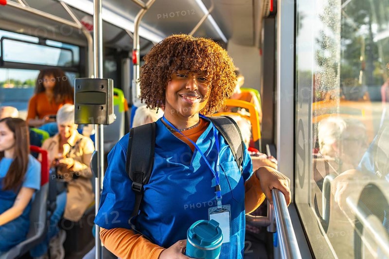 Young nurse in blue scrubs and backpack smiles at camera while standing on a city bus, holding a reusable cup during her commute as an essential healthcare worker