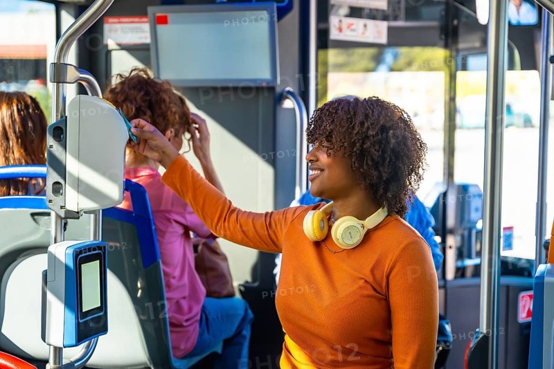 Smiling woman with headphones validating a contactless travel card on a modern electronic validator inside a city bus, commuting comfortably using public transit technology and routine