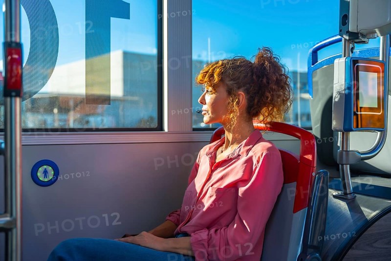 Woman seated on a city bus, gazing out the window in quiet thought during commute as sunlight highlights her curly hair and contemplative profile in the urban interior