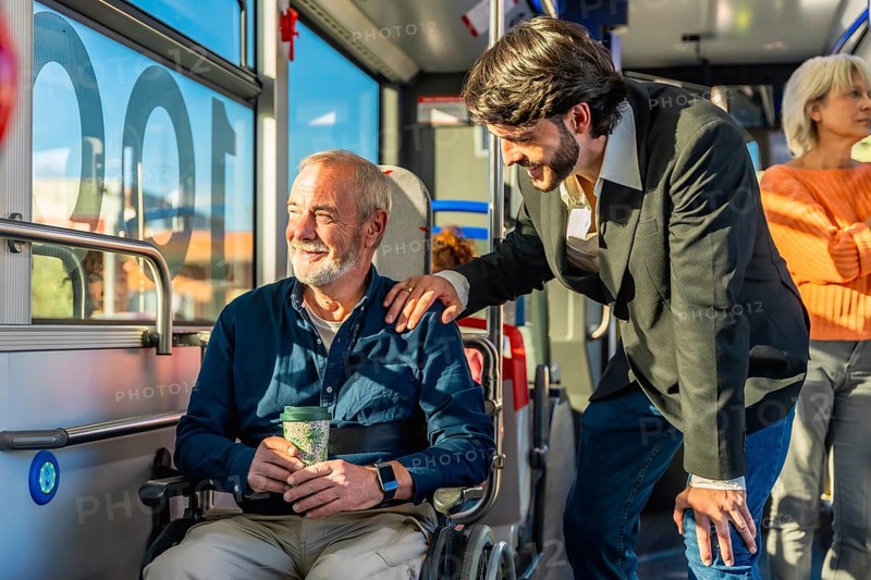 Young man offering support and kindness to a smiling senior passenger using a wheelchair, showcasing empathy and accessibility during their journey on a bus