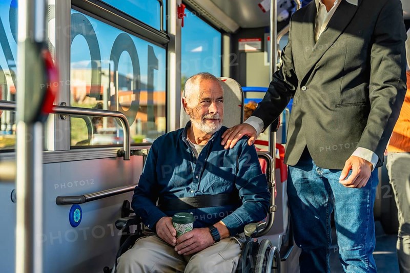 Senior man with a disability using public transportation, sitting in his wheelchair on a bus while receiving a helping hand and emotional support from a caring assistant