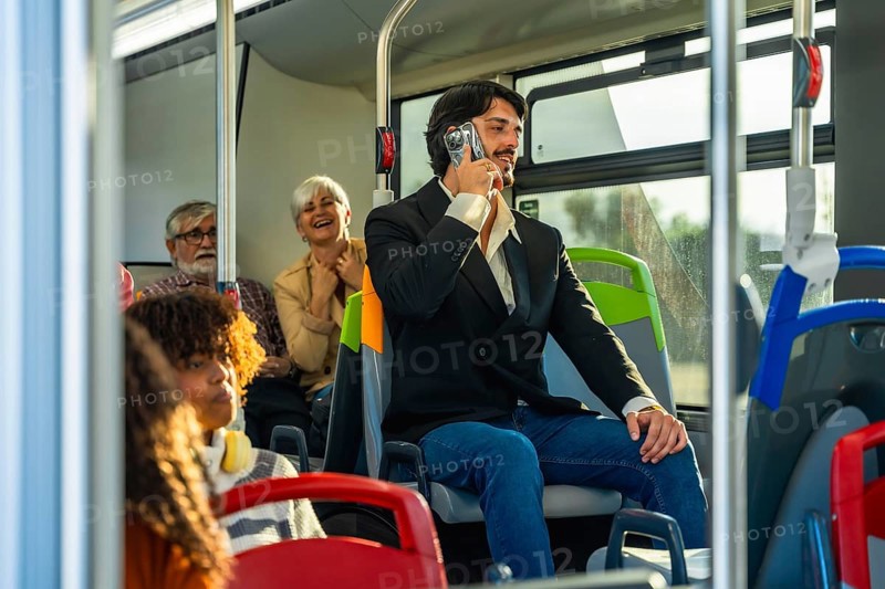 Man holding a vintage camcorder to his ear, pretending it is a cell phone, engaging in a playful conversation while other commuters are present on public urban transportation