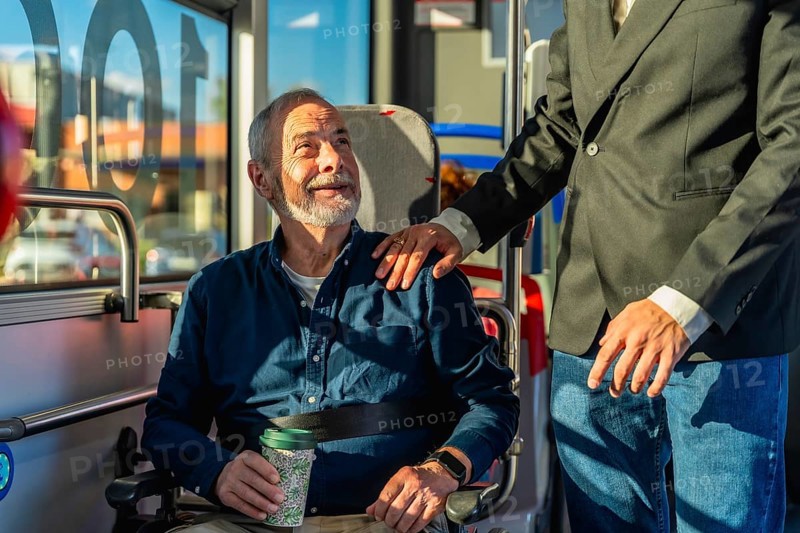 Young caregiver places hand on shoulder of smiling senior man in wheelchair during accessible public transport ride, sharing warmth, trust and comfortable mobility