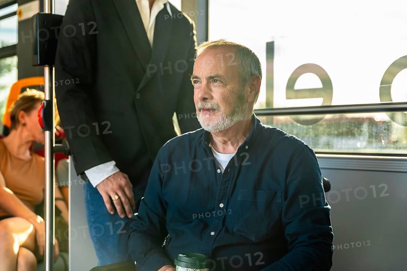 Senior man on a city bus, seated by the window with a reusable cup, gazing thoughtfully outside during a calm urban commute in daylight, relaxed and contemplative