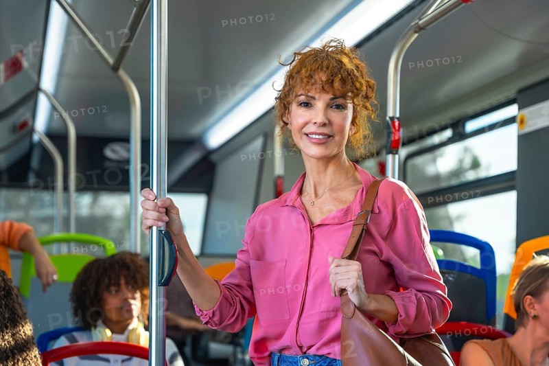 Smiling redhead woman standing and holding a pole on a busy city bus, commuting comfortably among passengers during a bright, everyday urban transit journey