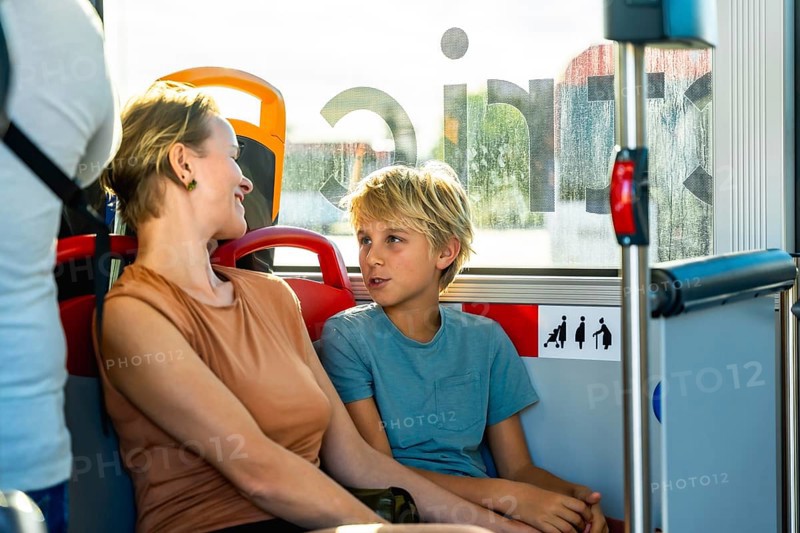 Mother and son smiling and chatting on a sunlit city bus during their commute, sharing a relaxed bonding moment as they travel together through the urban landscape
