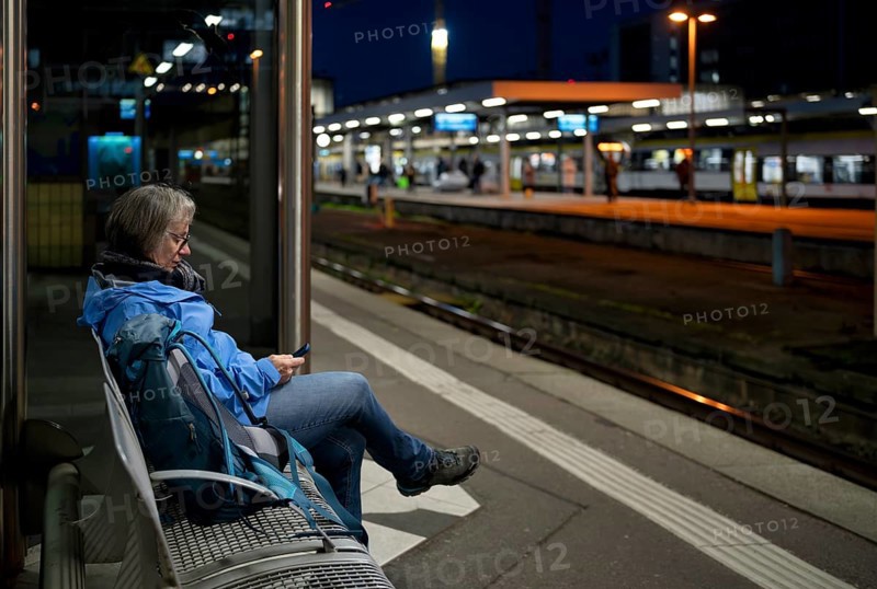 Night shot, elderly woman, traveler, sitting alone on a bench on a platform, backpack, using smartphone, cell phone, main train station, Stuttgart, Baden-Württemberg, Germany