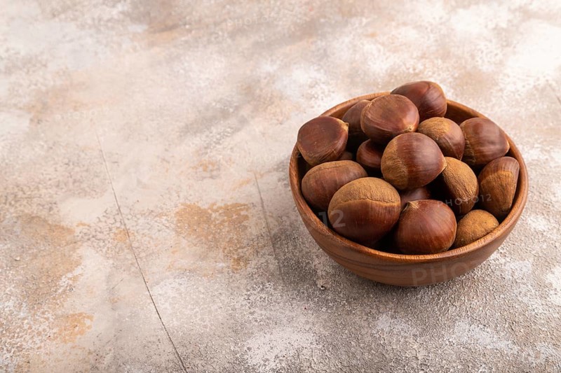 Wooden bowl with raw edible ?hestnuts on brown concrete background, side view, copy space, minimalism