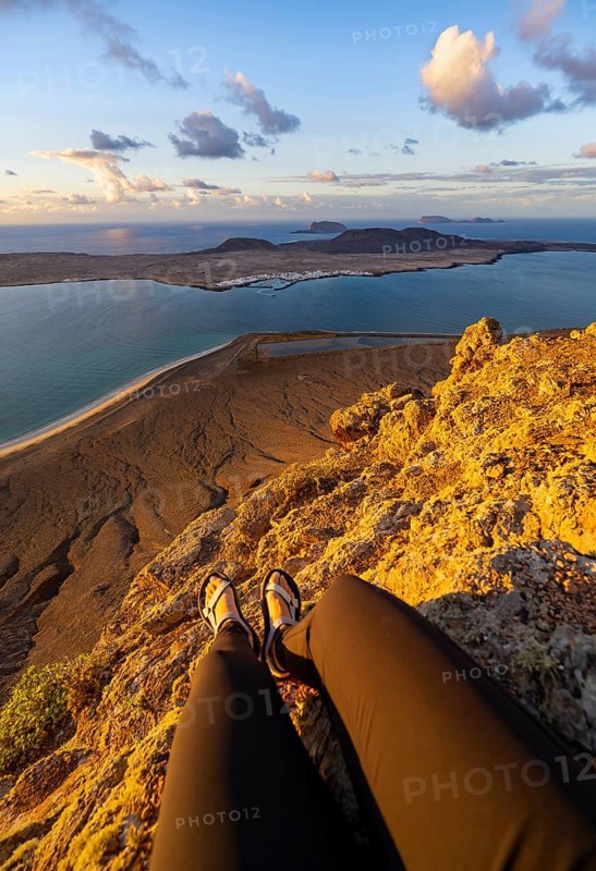 First person view, legs of a woman sitting on a steep cliff, view of the sea and the island of La Graciosa, Mirador del Porrito viewpoint at sunset, Lanzarote, Canary Islands, Spain
