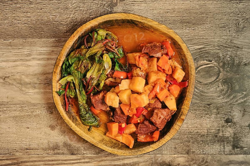 A warm bowl of beef stew features tender chunks of meat, potatoes, carrots, and bell peppers. Steamed greens salad leaves accompany the dish, highlighting flavors and colors in a cozy setting
