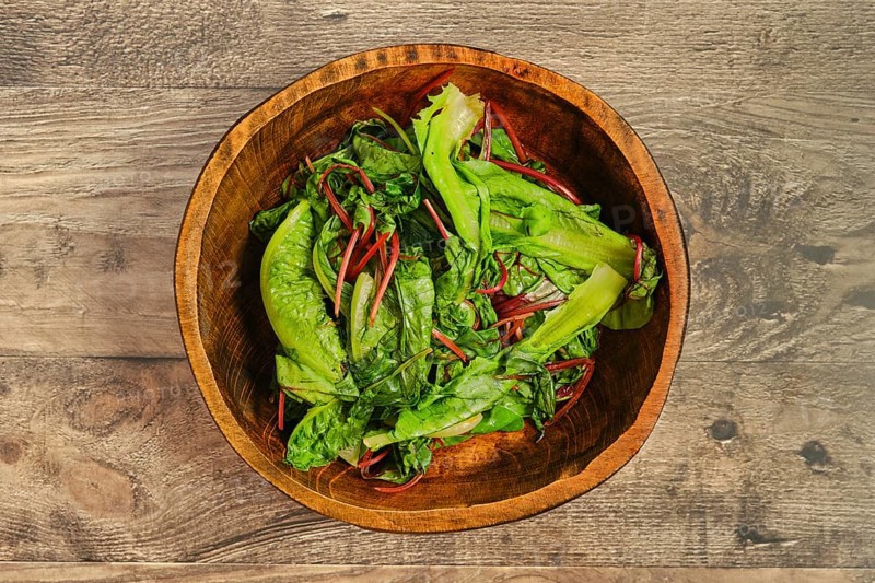 A wooden bowl filled with steamed greens on a rustic wooden tabletop. The leafy vegetables, including romaine and other greens, look crisp and healthy, ready for preparation