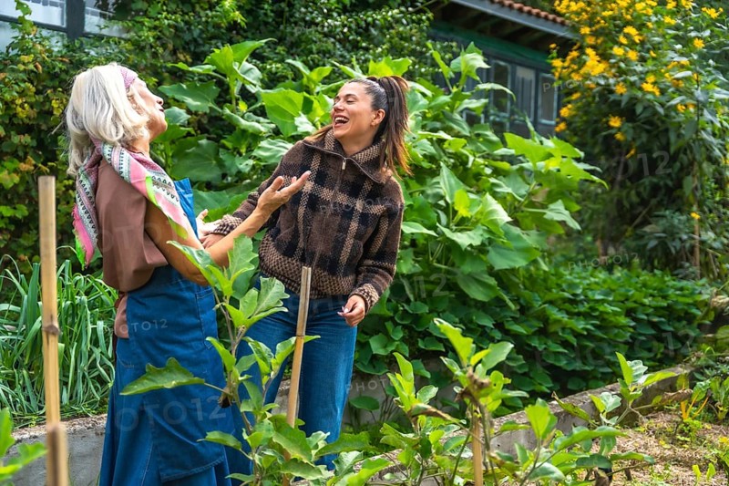 Two cheerful women of different generations are laughing and bonding outdoors, tending to plants in a vibrant home garden, sharing a joyful moment of connection and healthy lifestyle