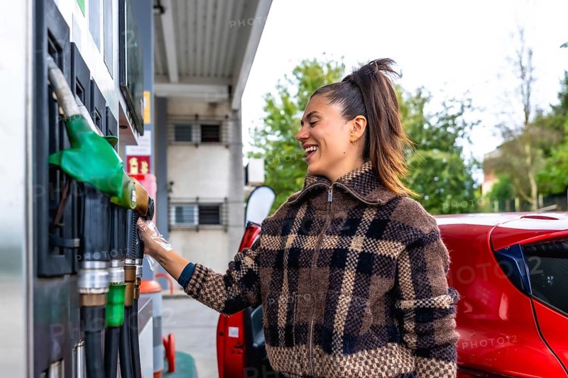 Woman smiling at a gas station while holding a fuel pump nozzle beside her red car in a plaid jacket, preparing to refuel on a sunny day during a road trip stop
