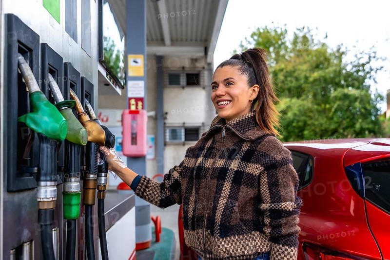 Woman smiling and wearing a protective glove while selecting a fuel pump to refuel her red car at a gas station, showcasing daily tasks and vehicle maintenance