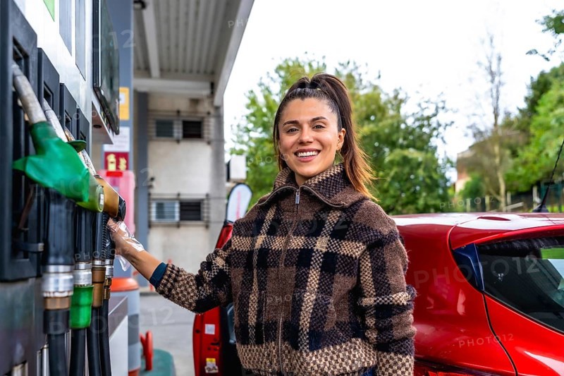 Smiling young woman in a plaid jacket and protective glove refuels her red car at an urban gas station, close up on fuel pump during daytime roadside stop