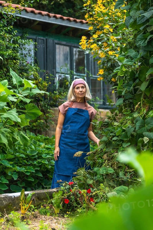 Senior woman in an apron and headband standing in a lush home vegetable garden, smiling while tending colorful flowers and green plants, enjoying a peaceful gardening hobby
