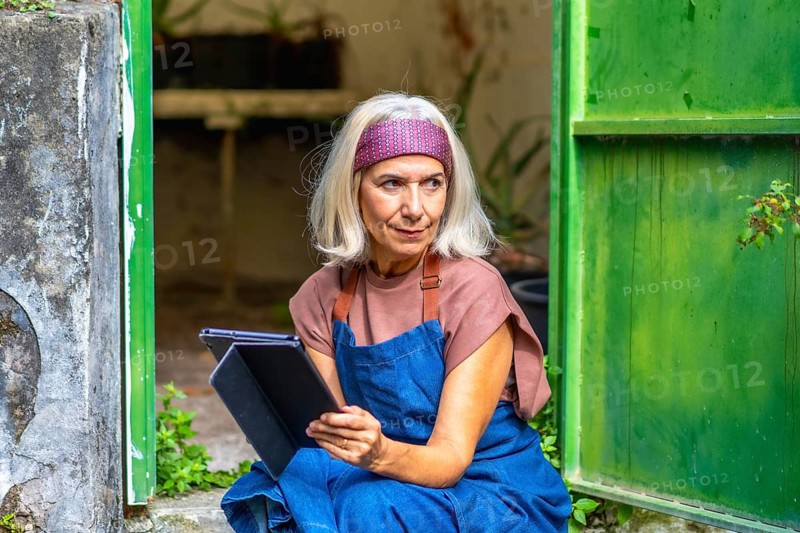 Senior woman artist in an apron and headband working on a digital tablet outside a rustic green building, thoughtful and focused as she sketches and plans her creative project