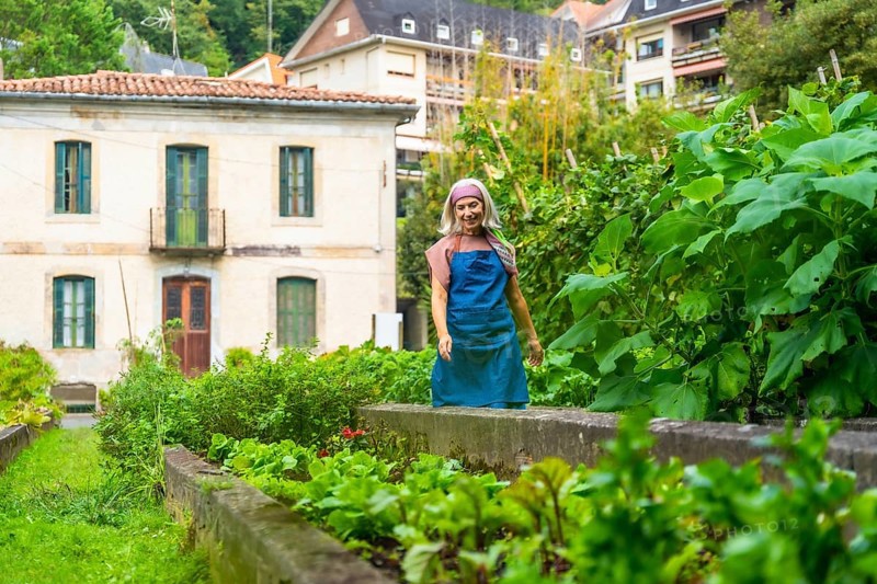 Woman gardener smiling while standing in a lush, rustic raised bed vegetable garden, cultivating fresh produce with green plants and residential buildings in the background